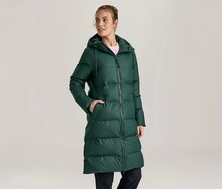 Femme portant un long manteau matelassé vert foncé à capuche, une main dans la poche, regardant sur le côté.