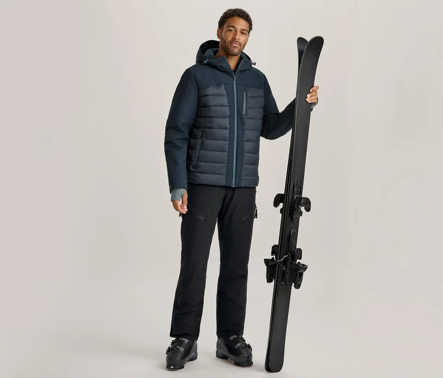 Homme en doudoune bleu foncé, pantalon et chaussures de ski noirs, tenant une paire de skis noirs à la verticale.