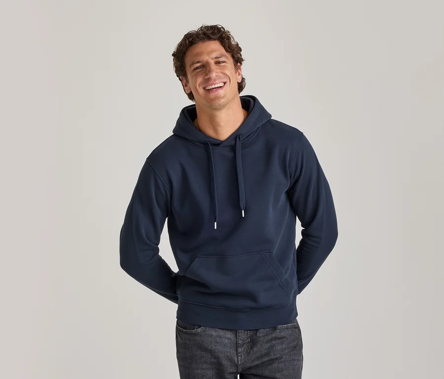 Homme souriant portant un sweat à capuche bleu marine avec des cordons blancs et un jean foncé, les mains derrière le dos.