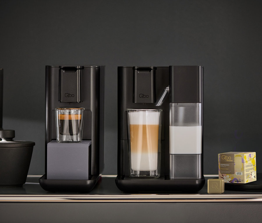 Deux machines à café Qbo noires sur une étagère sombre. La machine de gauche contient un verre d'espresso. La Qbo MILK MASTER Velvet Black de droite contient un verre de latte macchiato en couches et un réservoir à lait transparent. À droite se trouvent une boîte jaune de capsules d'espresso Qbo et une capsule dorée.
