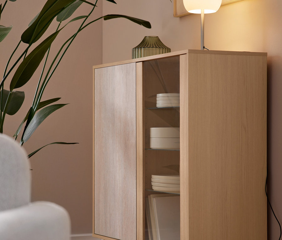 Un meuble en bois clair avec une porte pleine et une porte vitrée repose sur un parquet. À l'intérieur de la section vitrée se trouvent des assiettes et des bols blancs empilés. Sur le meuble, une lampe sphérique blanche allumée et un vase vert côtelé sont placés à côté d'une grande plante verte. Une œuvre d'art abstraite encadrée est accrochée au mur marron clair au-dessus de la lampe, et un fauteuil blanc est partiellement visible à gauche.