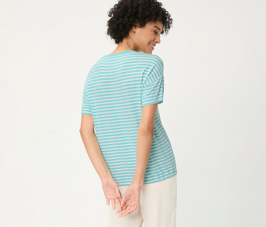 Femme de dos, regardant par-dessus son épaule droite et souriant, portant un T-shirt Olsen rayé turquoise et gris clair et un pantalon clair, les mains jointes derrière le dos.