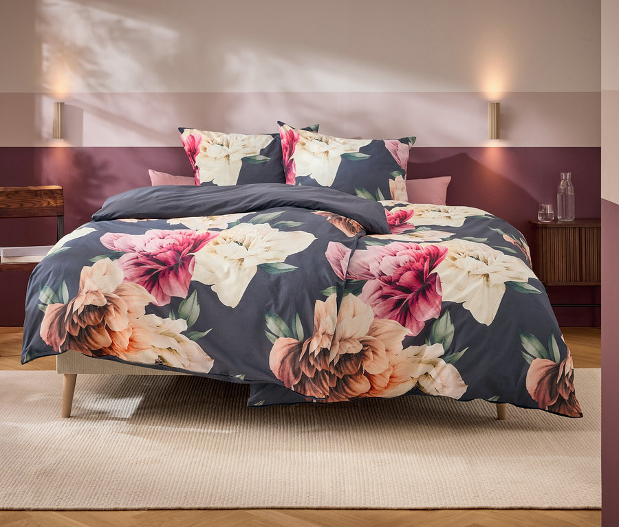Un lit avec une parure de lit gris foncé à grands motifs floraux roses, blancs et oranges se trouve dans une chambre aux murs bicolores et aux meubles en bois.