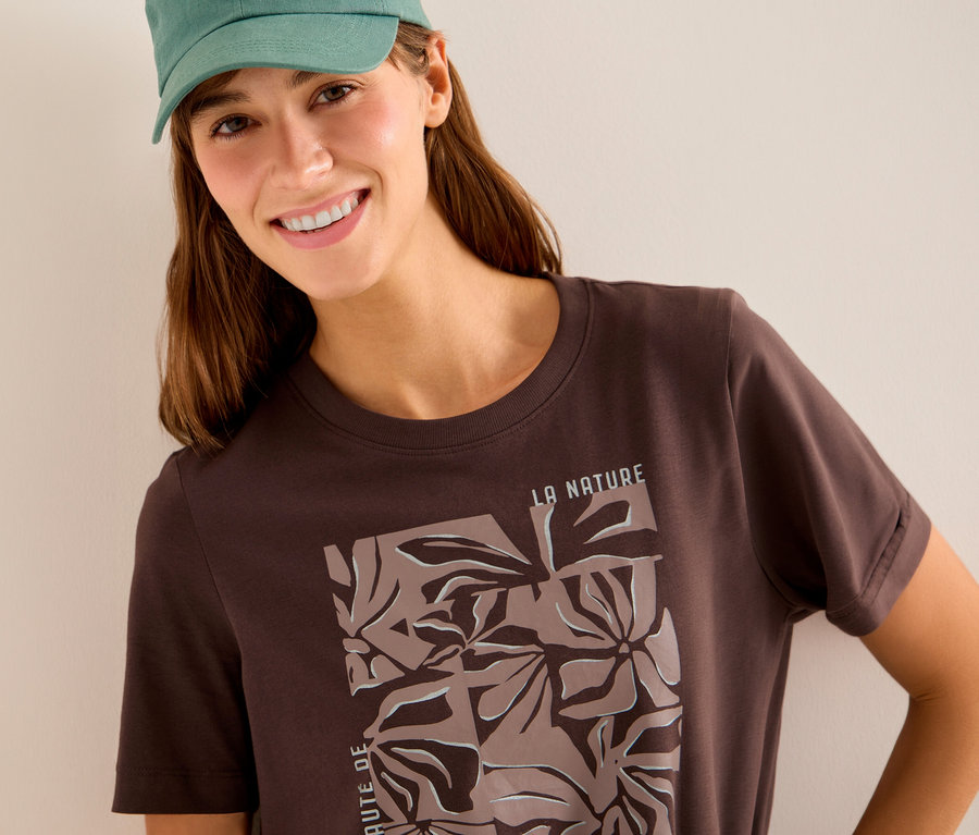 Femme souriante portant une casquette verte et un T-shirt à motif CECIL marron foncé avec un motif floral.
