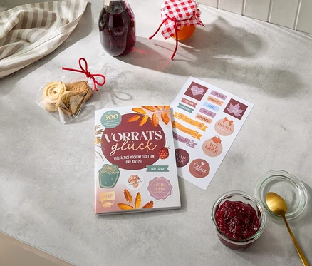 Sur un comptoir gris, on trouve un torchon rayé, un sachet de biscuits avec un ruban rouge, une bouteille de liquide rouge foncé et un pot de confiture. À côté, un livre intitulé « Vorratsglück » avec « Vielfältige Küchenetiketten und Rezepte » et une feuille d'autocollants avec des phrases comme « Für dich » et « Homemade ». En bas à droite, un pot ouvert de confiture rouge avec une cuillère dorée.