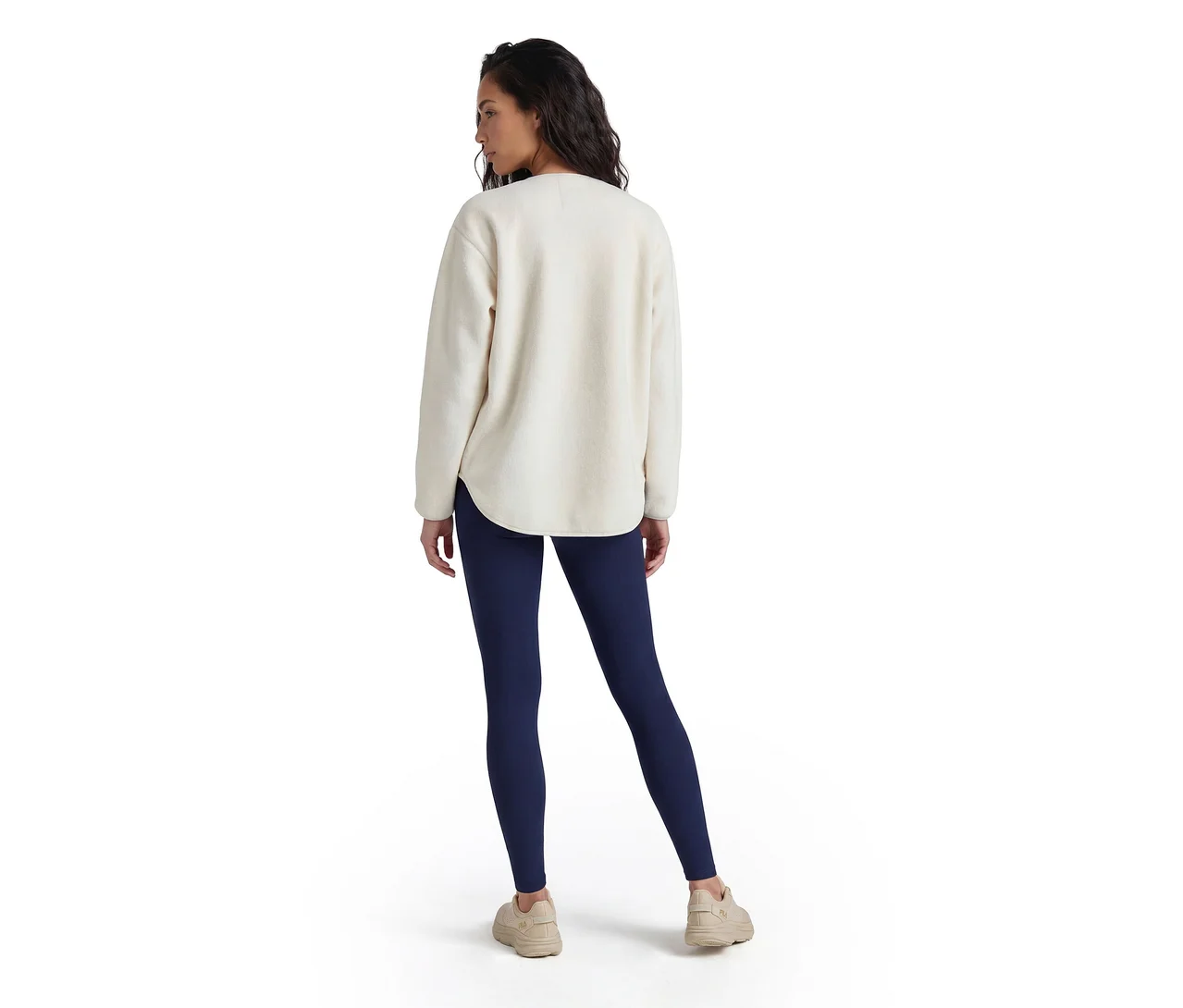 Femme vue de dos, portant un haut à manches longues de couleur crème, un legging bleu foncé et des baskets beige clair, regardant vers sa droite.