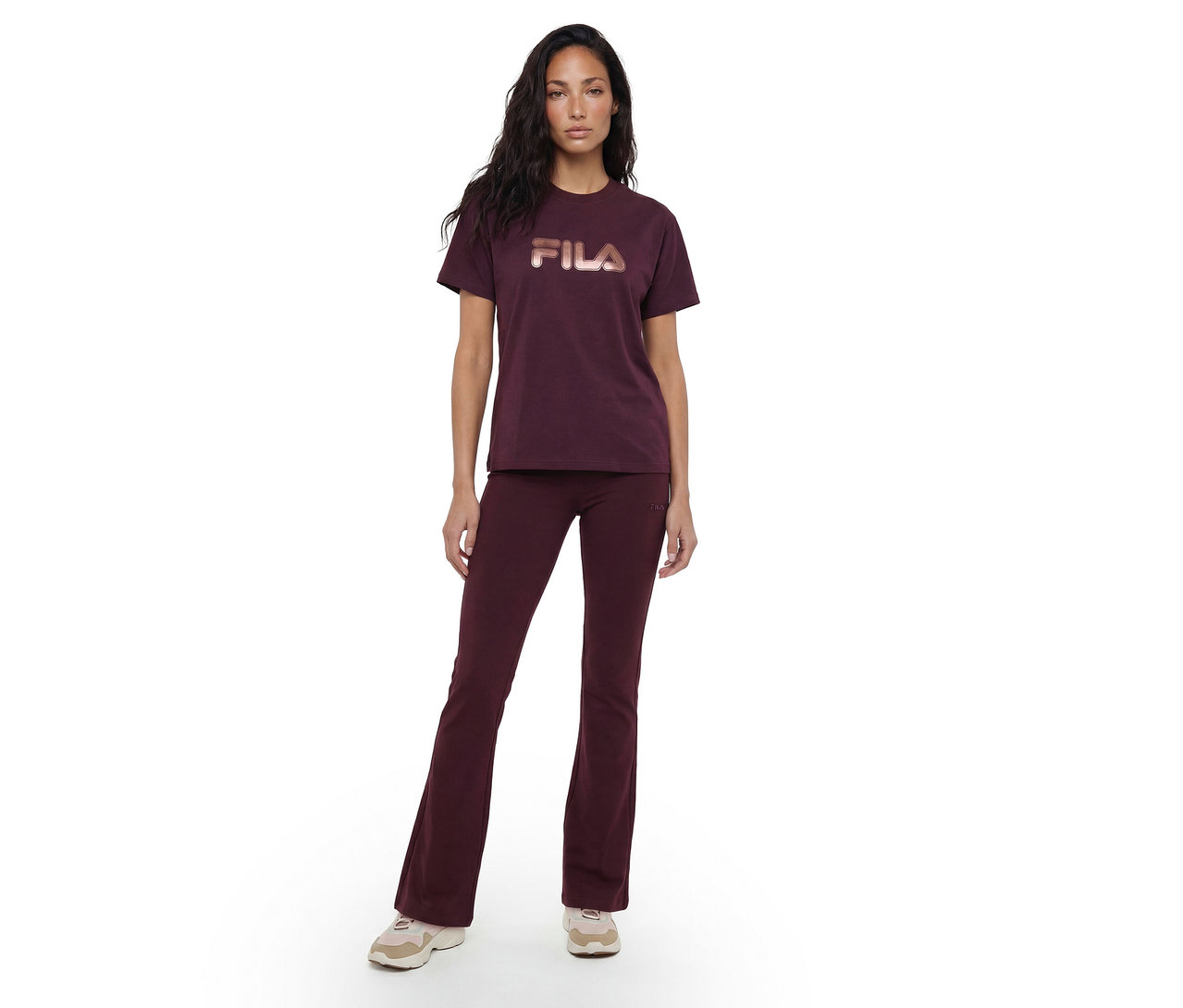 Une femme se tient de face, vêtue d'un t-shirt rouge foncé avec un logo "FILA" couleur or rose et d'un pantalon évasé rouge foncé assorti, associé à des baskets de couleur claire.