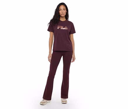 Une femme se tient de face, vêtue d'un t-shirt rouge foncé avec un logo "FILA" couleur or rose et d'un pantalon évasé rouge foncé assorti, associé à des baskets de couleur claire.