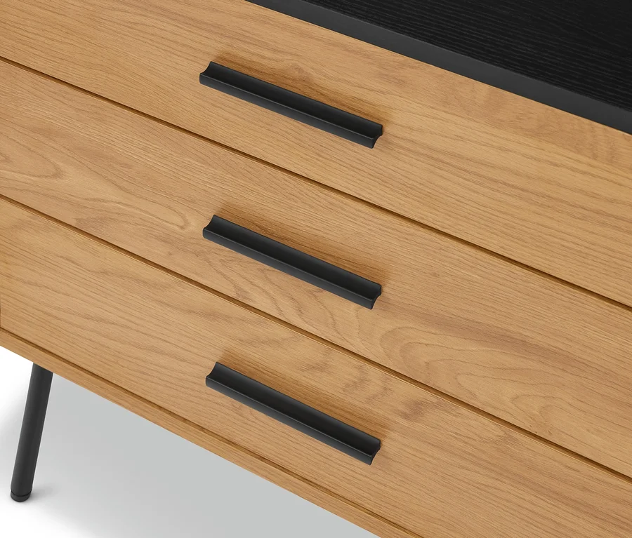 Gros plan d'une commode en bois clair avec trois tiroirs et des poignées noires. Le plateau est noir, un pied noir est visible à gauche.