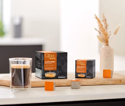 Deux boîtes de capsules Qbo Caffè MINAS VERDE, un verre de café et des capsules individuelles sur une planche en bois sur un comptoir de cuisine, avec des biscuits et des fleurs séchées.