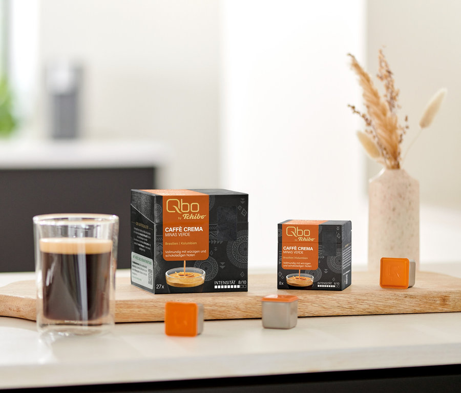 Deux boîtes de capsules Qbo Caffè MINAS VERDE, un verre de café et des capsules individuelles sur une planche en bois sur un comptoir de cuisine, avec des biscuits et des fleurs séchées.