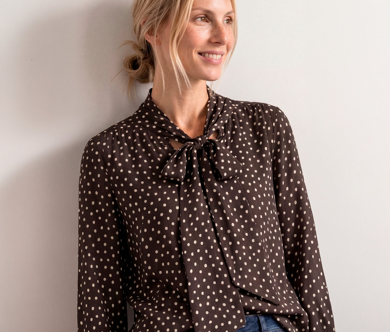 Femme souriante aux cheveux blonds attachés, portant une blouse Street One marron foncé à pois blancs avec un nœud au col, appuyée contre un mur blanc.