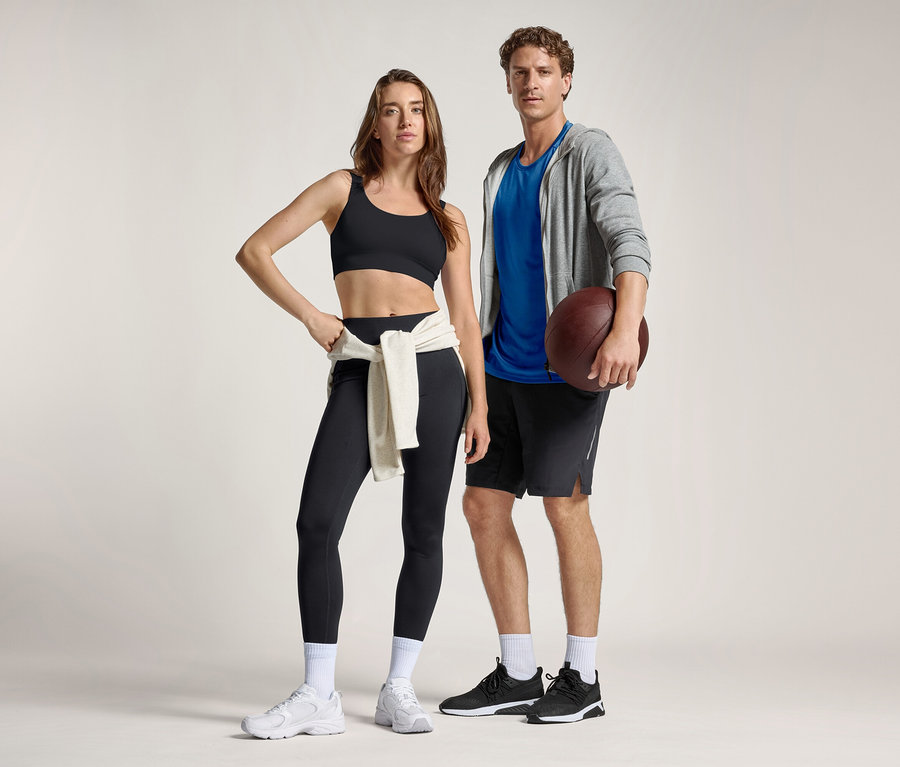 Une femme en brassière de sport noire et leggings de sport « ActivePower » avec un sweat-shirt noué à la taille, se tient à côté d'un homme en t-shirt bleu, sweat à capuche gris et short, tenant un ballon de football américain.