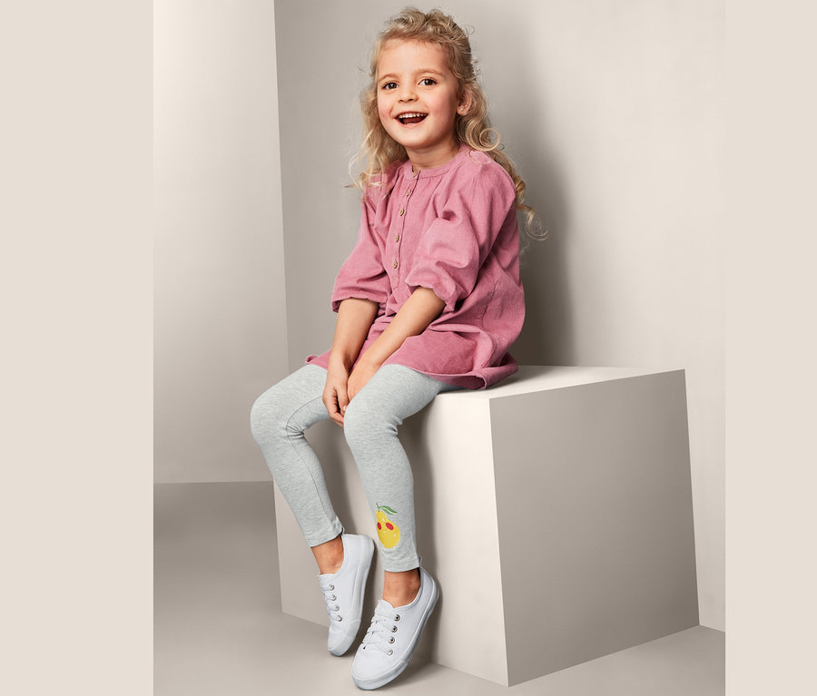 Une fillette blonde souriante est assise sur un cube blanc et porte une robe rose et 2 leggings avec un imprimé citron et des baskets blanches.