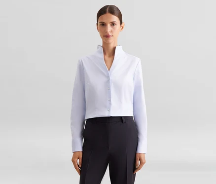 Femme portant une chemise courte bleu clair à col montant et un pantalon foncé, posant devant un fond blanc.