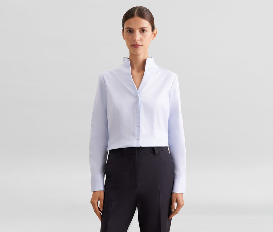 Femme portant une chemise courte bleu clair à col montant et un pantalon foncé, posant devant un fond blanc.