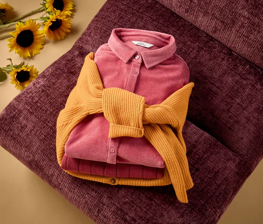 Une chemise en velours côtelé rose pliée et un haut côtelé rose foncé, avec un cardigan en mérinos jaune noué autour, sont posés sur une surface texturée rouge foncé. Des tournesols sont visibles en arrière-plan.