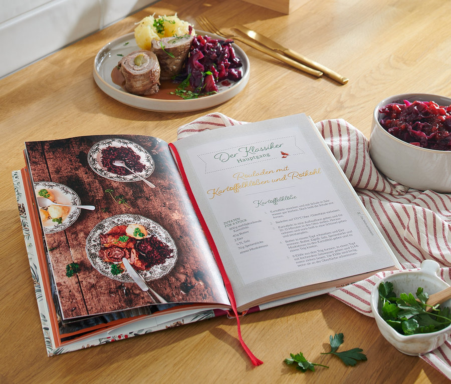 Livre de cuisine ouvert sur une table en bois avec des aliments dans des bols et sur une assiette, à côté le livre « Christmas is a feeling » (en allemand).