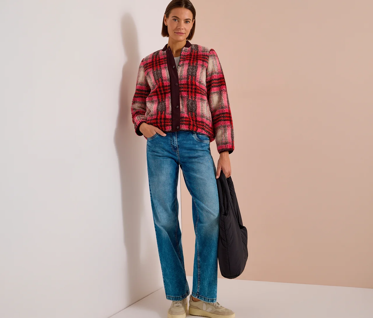 Une femme aux cheveux courts et bruns est debout, portant un blouson CECIL à carreaux rouge, rose et beige, un jean bleu et des baskets claires, tenant un sac noir.
