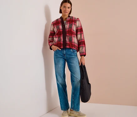 Une femme aux cheveux courts et bruns est debout, portant un blouson CECIL à carreaux rouge, rose et beige, un jean bleu et des baskets claires, tenant un sac noir.