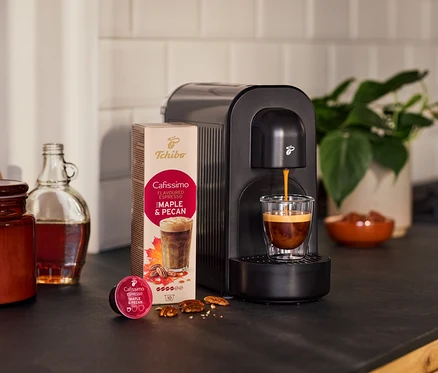 Machine à café noire préparant un espresso dans un verre. À côté, un paquet de capsules saveur érable-pécan, une capsule, des noix, une bouteille et une plante.