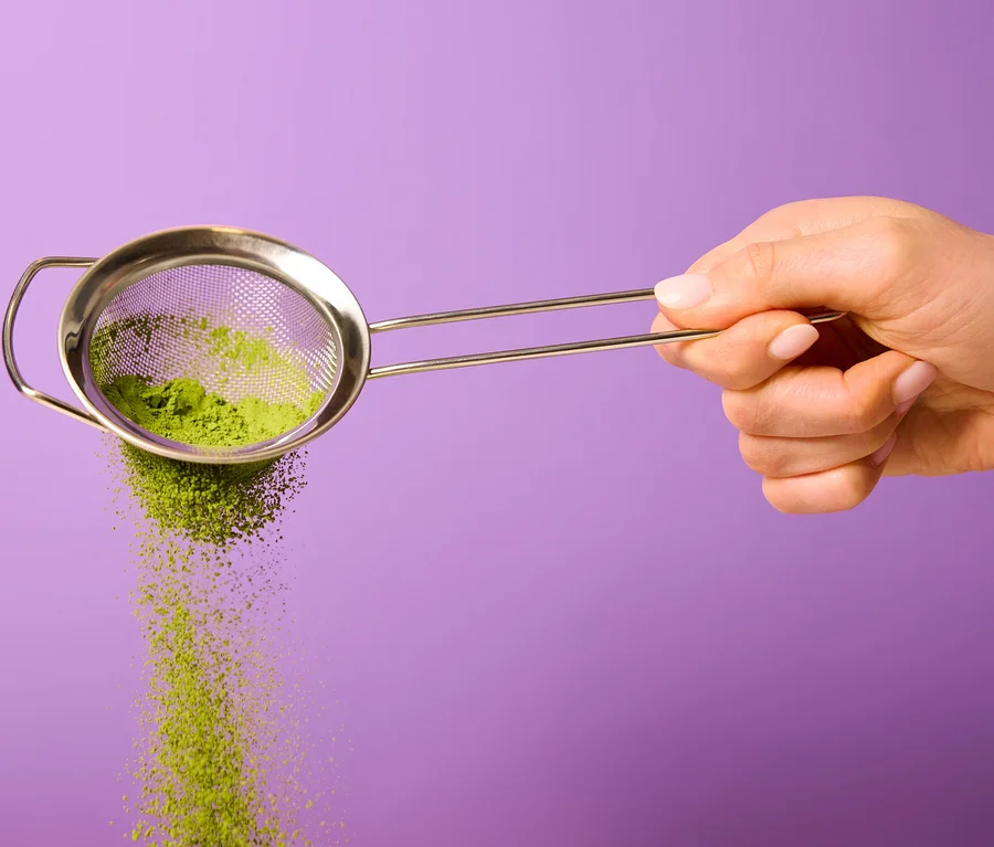 Une main tient une passoire métallique d'où tombe de la poudre de Bio Matcha by Tchibo verte sur un fond violet.