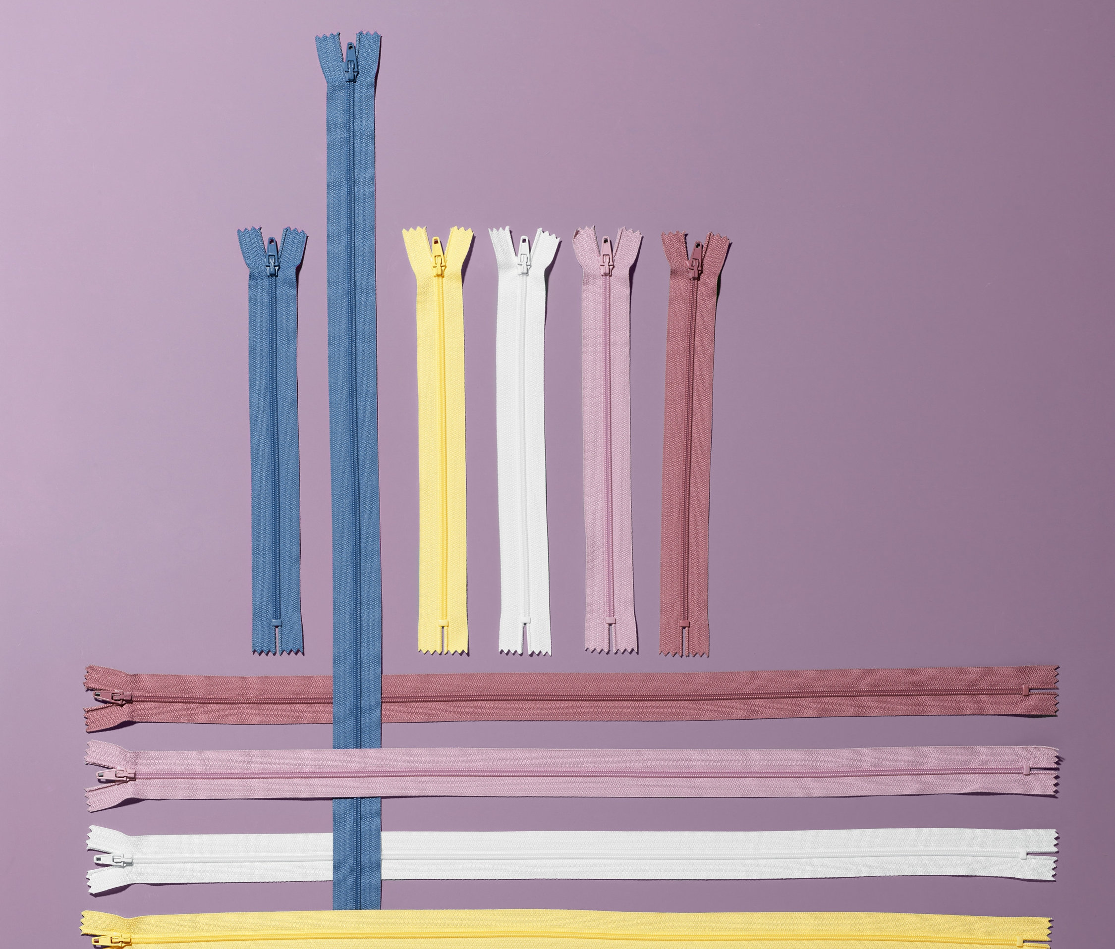 Sur un fond violet, se trouvent 10 fermetures éclair de différentes couleurs et longueurs.