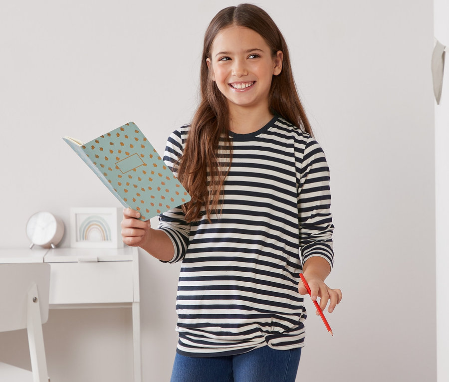 Une fille tient un cahier ouvert et un crayon rouge. Elle porte un t-shirt manches longues avec nœud décoratif bleu-blanc à rayures.