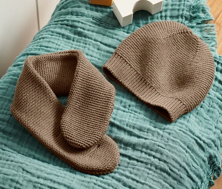 Un bonnet et une écharpe en maille marron sont posés sur un tissu bleu-vert sur un tabouret en bois, à côté de cubes blancs et jaunes.