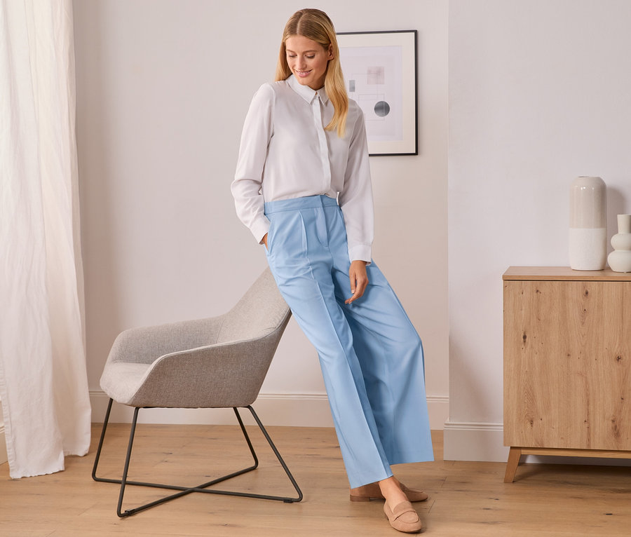 Une femme portant une chemise blanche et un pantalon tissé bleu clair s'appuie contre une chaise grise.