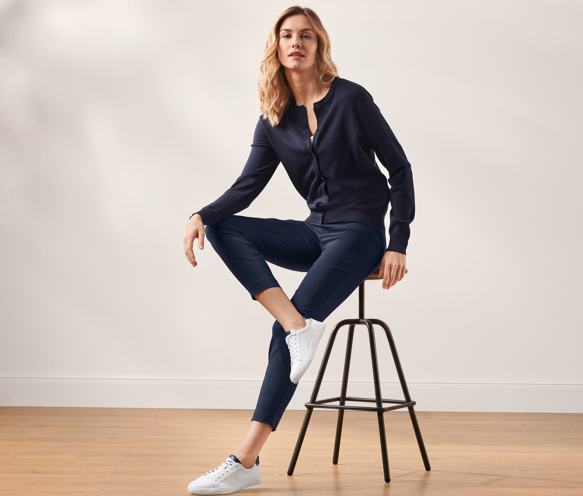 Une femme blonde est assise sur un tabouret et porte un pantalon stretch bleu foncé et un cardigan.