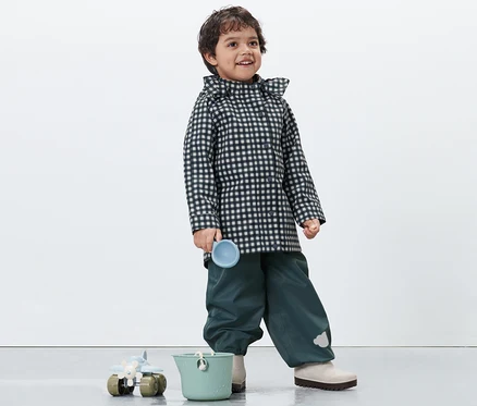 Un garçon portant un imperméable pour tout petit à carreaux tient une tasse bleue. Un seau vert et un jouet sont en bas.