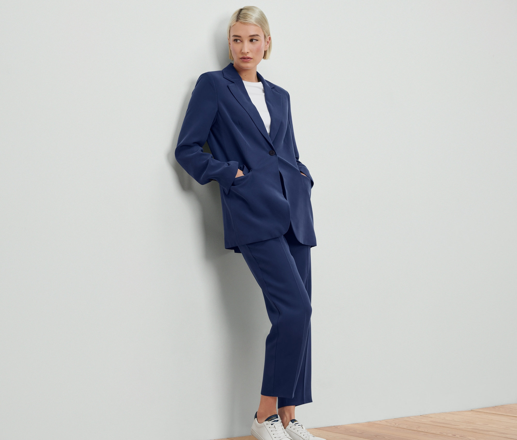 Une femme appuyée contre un mur porte un blazer en toile bleu et un pantalon assorti.