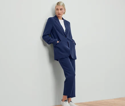 Une femme appuyée contre un mur porte un blazer en toile bleu et un pantalon assorti.