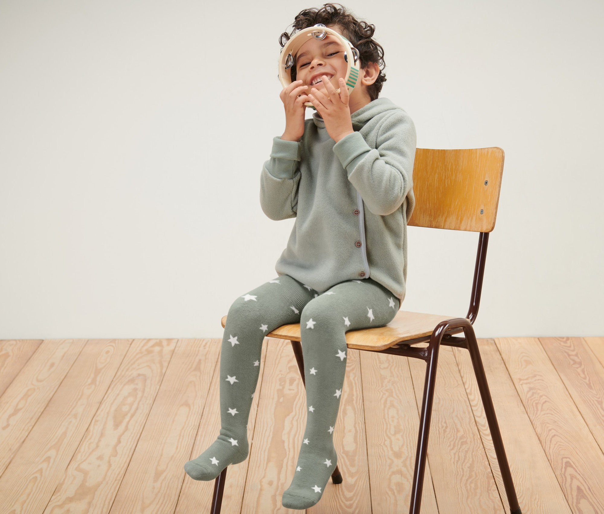 Un garçon est assis sur une chaise et joue avec un tambourin. Il porte des collants thermiques bleus et verts pour enfants.