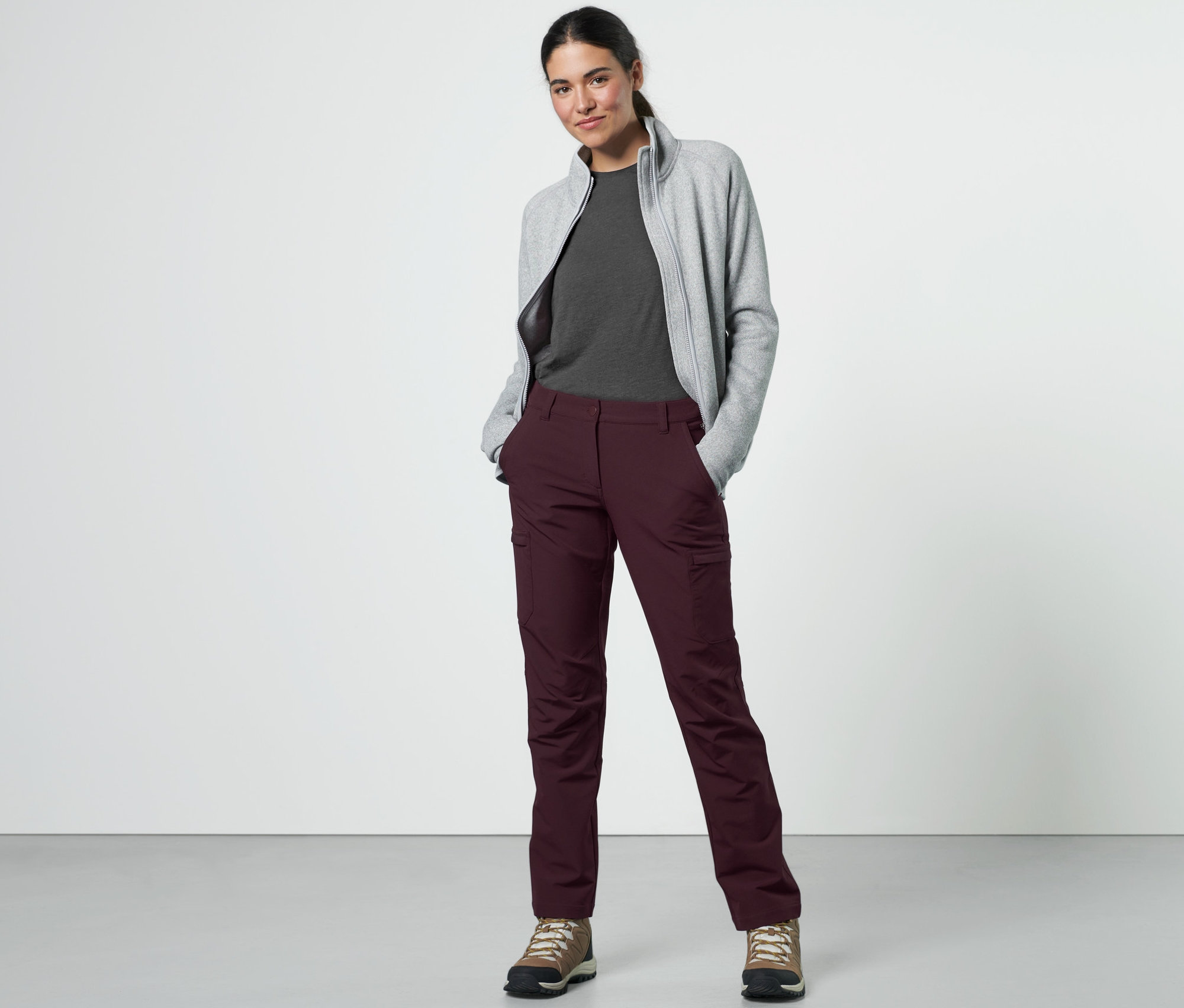 Une femme pose en pantalon cargo softshell bordeaux, t-shirt gris et sweat-shirt zippé gris.