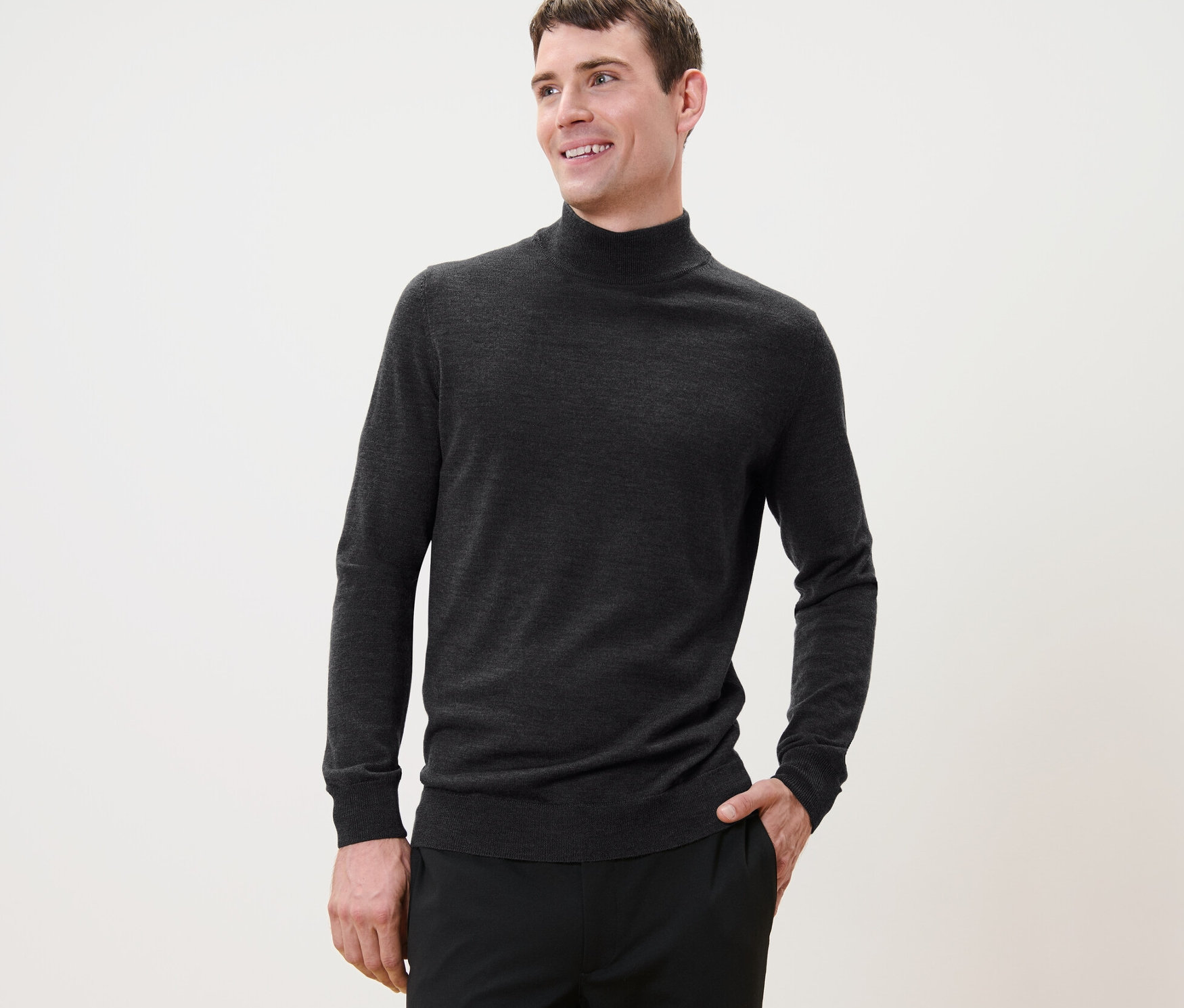 Un homme aux cheveux courts pose, portant un pull en laine mérinos anthracite et un pantalon noir.