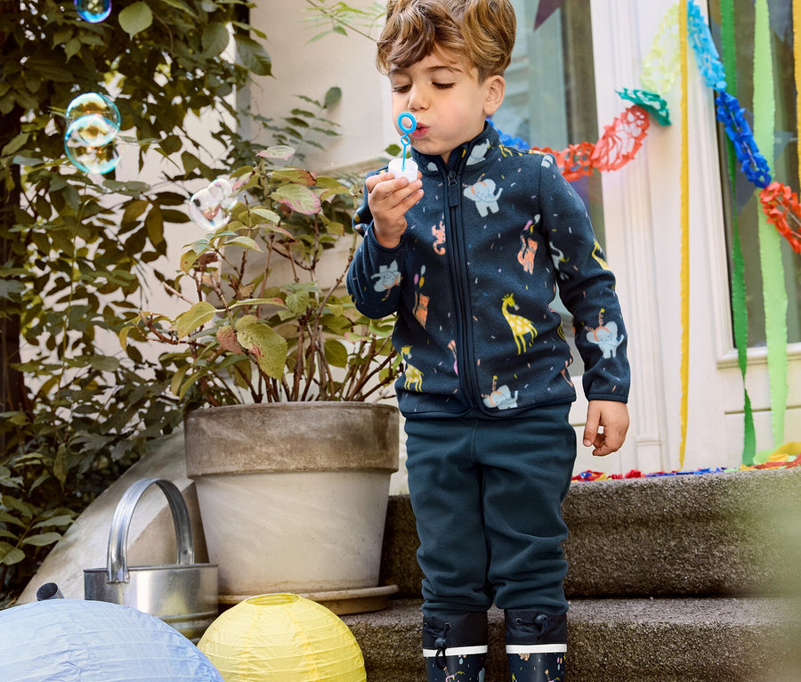 Garçon faisant des bulles, portant une jaquette en polaire, un pantalon en polaire et des bottes de pluie pour enfant.