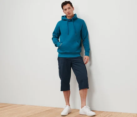 Un homme appuyé contre un mur porte un sweat à capuche bleu-vert et un bermuda cargo 3/4 bleu marine.