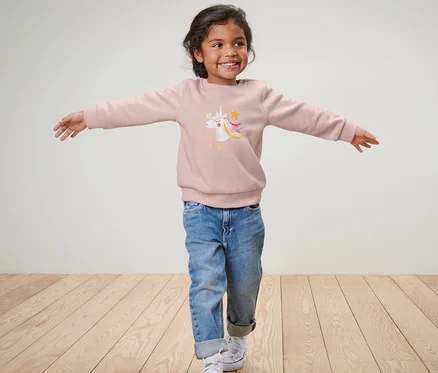 Une fille aux bras écartés porte un sweater pour enfant licorne, un jean bleu et des baskets.