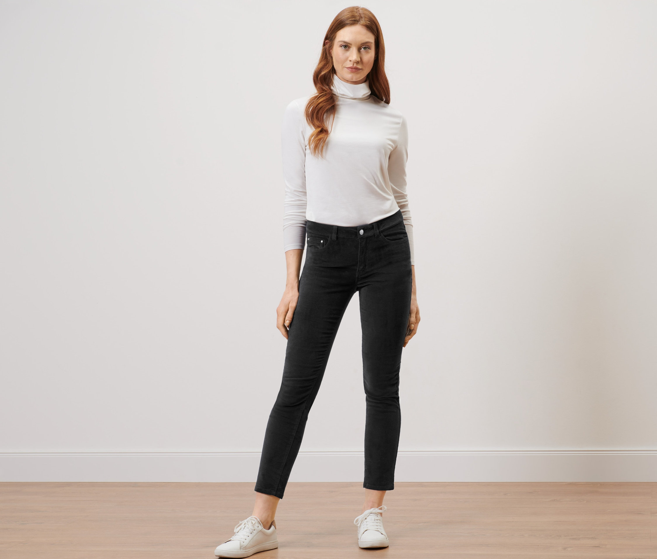 Une femme debout sur un plancher en bois porte un pantalon en velours noir et un col roulé blanc.