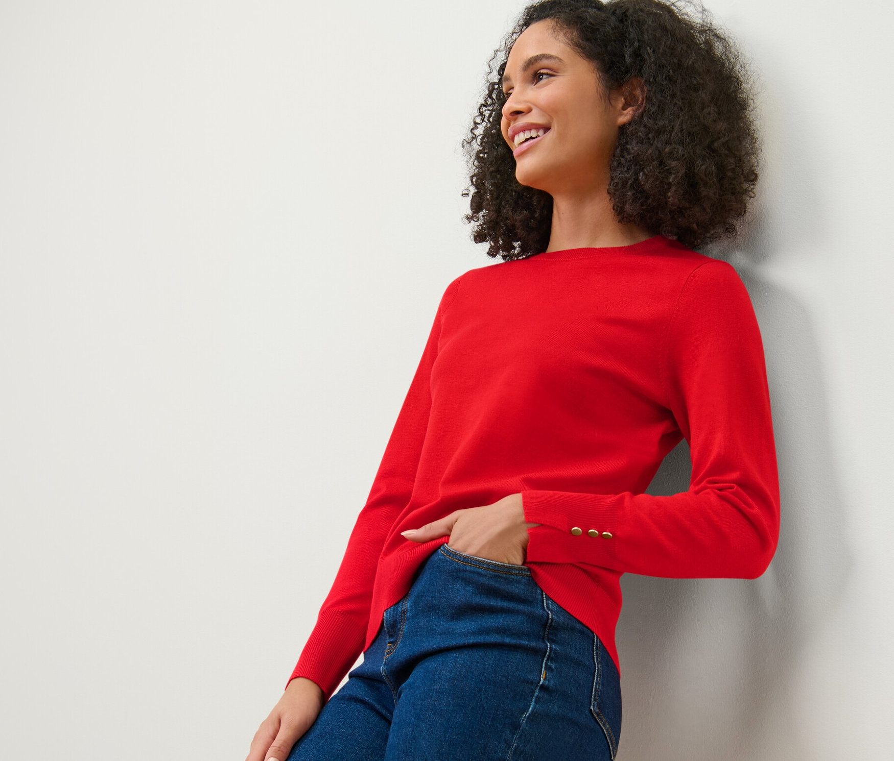 Femme souriante appuyée contre un mur portant un pull en maille fine rouge et un jean.