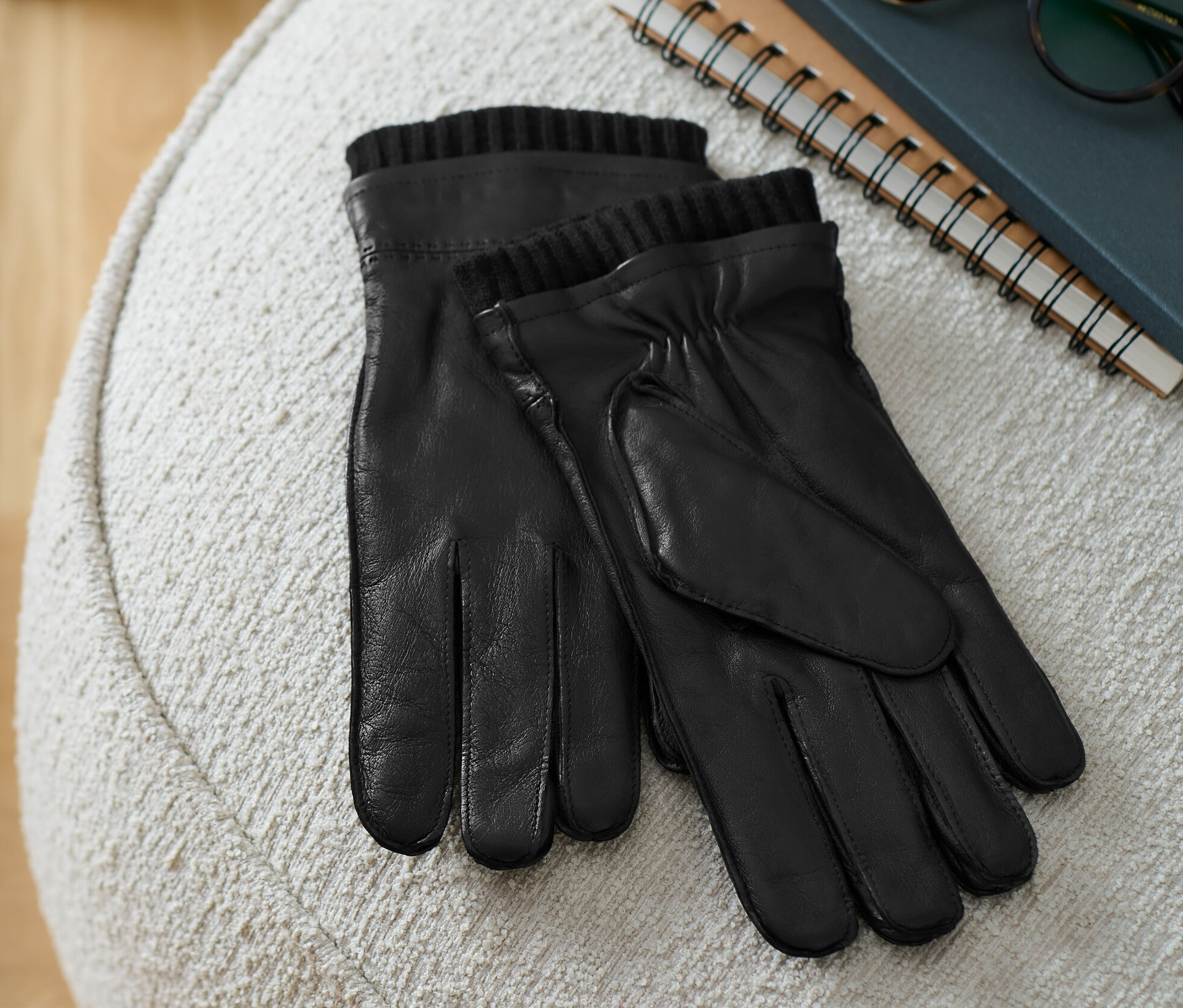 Des gants en cuir noirs sont posés sur un tabouret beige à côté d'un livre et de lunettes.