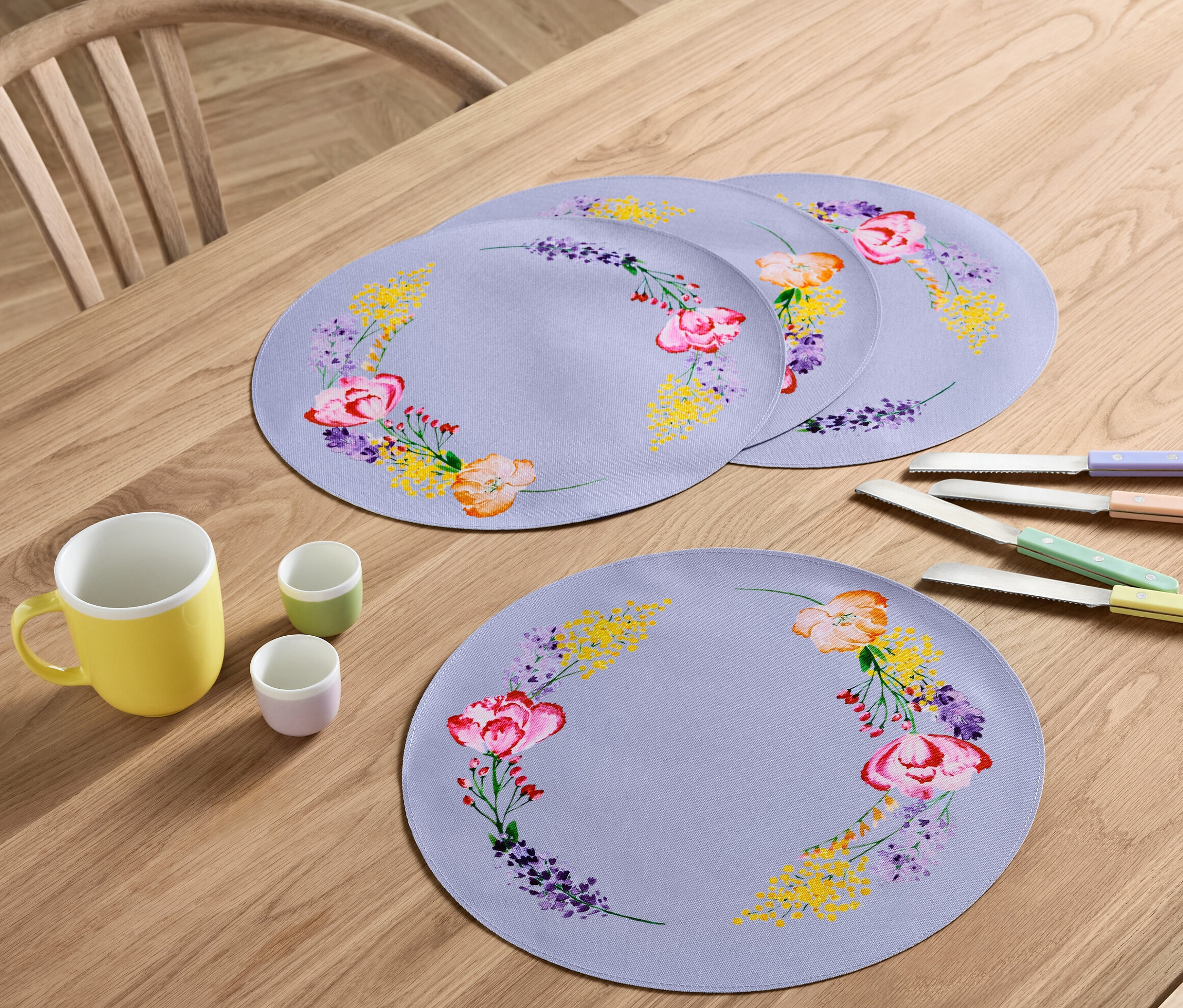 Sur une table en bois sont disposés quatre sets de table à motifs floraux, une tasse jaune, deux petites tasses et quatre couteaux avec des manches colorés.