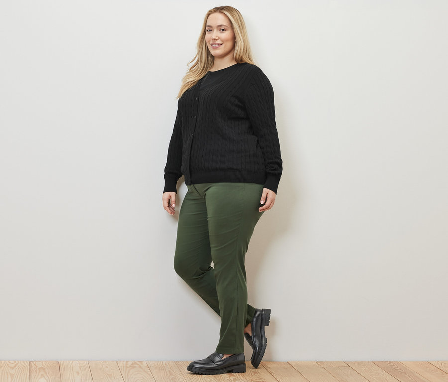 Une femme pose en cardigan en maille fine noire à motif torsadé et un pantalon en bengaline vert.