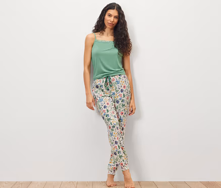 Une femme debout devant un mur blanc porte un haut vert et un pantalon de pyjama.
