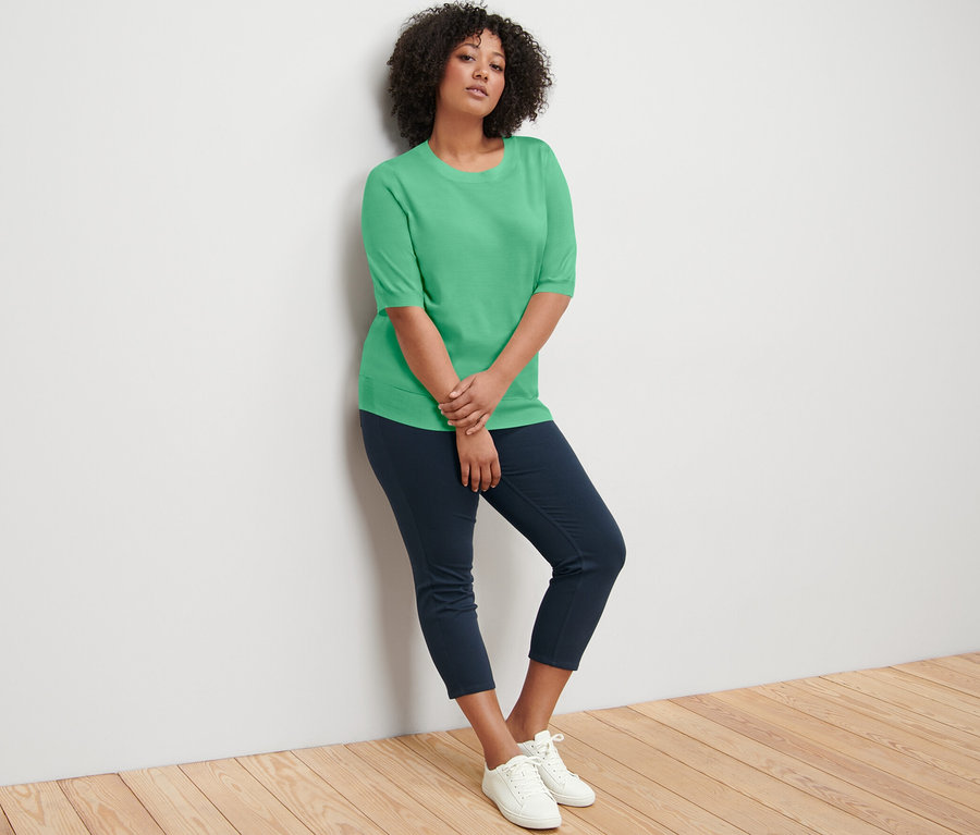 Une femme appuyée contre un mur porte un pull en maille fine vert, un pantalon bleu marine et des baskets blanches.