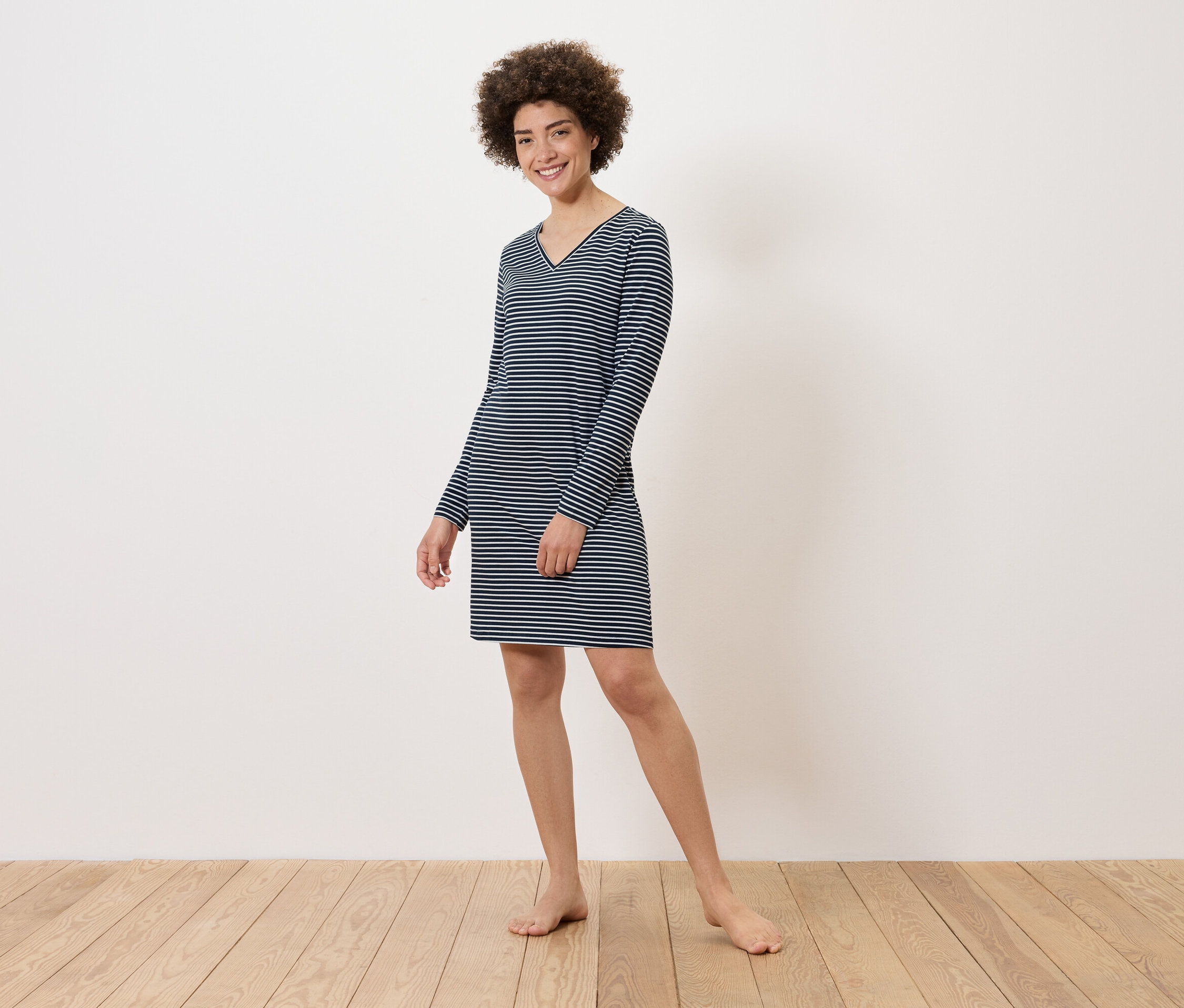 Une femme se tient pieds nus sur un plancher en bois et porte une robe rayée à manches longues.