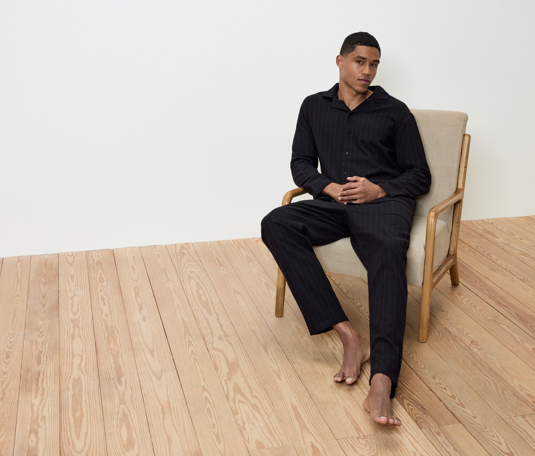 Un homme est assis pieds nus dans un fauteuil beige sur un plancher en bois, vêtu d'un pyjama rayé noir.
