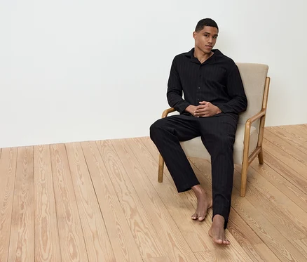 Un homme est assis pieds nus dans un fauteuil beige sur un plancher en bois, vêtu d'un pyjama rayé noir.